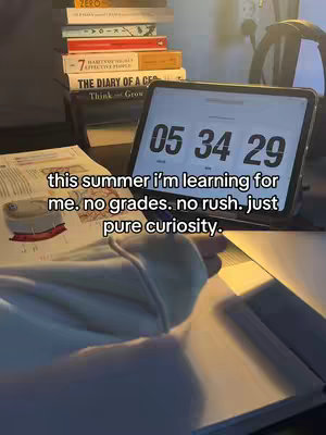 A close-up shot shows a desk with various items. A stack of books is on the left side of the frame, slightly out of focus, with titles like 'The Diary of a CEO' visible. In the center, a tablet displays a digital timer with the time 05:34:29. The focus seems to be on a person's hands and the top of a notebook. The lighting is warm, and the overall feel is relaxed and studious.
