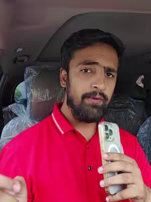 A man wearing a red polo shirt is holding a smartphone in a vehicle setting. He is looking at the camera with a semi-serious expression, while the background is of a car interior with seats and a windshield visible.