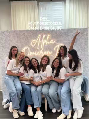 A picture of nine young women posing for a photo. They're all in t-shirts and jeans, smiling and embracing each other. Behind them is a white wall, and a neon sign that says Alpha Omicron Pi, indicating a sorority. There is a window in the background. The text overlay on the image says "pov: your house joins a Greek Bible Study."