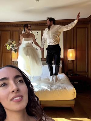 A young woman with dark hair and a thoughtful expression is partially visible in the bottom left corner. Behind her, a couple, the bride and groom, are jumping on a bed. The bride is wearing a white strapless gown and a veil. The groom is in a white shirt, dark pants, and white shoes. They are in a luxuriously decorated room with wood paneling and gold accent lamps. Black silhouette bird decorations are on the wall.