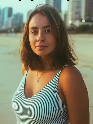 A woman with brown hair and a striped top is shown, with buildings and the beach visible in the background. The image has a warm, natural color palette.