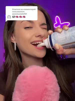 A woman is shown from the chest up, angled slightly to the left, with eyes closed and mouth open, in the act of consuming whipped cream, directly from a can and smiling. The background includes a pink fluffy prop and a neon purple butterfly, and a comment overlay.