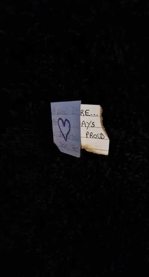 The image is a close-up on two torn pieces of paper against a completely black background. The piece on the left is a lighter blue and features a hand-drawn heart in blue pen above the words "love you". The right piece is off-white, has frayed edges, and the words "ARE...AYS PROUD". The darkness of the background emphasizes the contrast and isolates the written message.