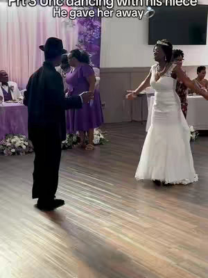 A candid shot from a wedding reception, showing a man in a black suit and a black hat dancing with a woman in a white wedding dress. Other guests are in the background, and the setting appears to be an event space, with tables and decorations visible.