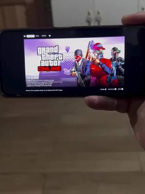A person holding a smartphone displaying the Grand Theft Auto Online game interface. The screen shows the game's title and characters. A hand is visible holding the phone against a background of wooden flooring and a blurry backdrop. The game graphics are vibrant and colorful.