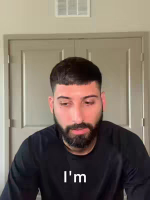 A man with a beard is looking down, wearing a black shirt with white text 'I'm' on it, seemingly in a bedroom.