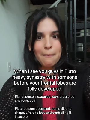 A selfie-style shot of a woman against an out-of-focus indoor backdrop. The woman has dark hair and fair skin, and is looking toward the camera with a slight expression. Text overlays provide context about astrology, specifically heavy synastry with Pluto. Red clothing is visible beneath a dark blazer.