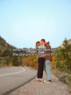 A couple kissing and embracing on a gravelly area adjacent to a road, backed by a forest and a partly cloudy sky. The woman has blonde hair and is wearing a light blue shirt and dark pants with white shoes. The man has short, dark hair and is wearing a brown zip-up hoodie, blue jeans, and dark-colored sneakers. A road curves alongside them, with a mountain in the backdrop under a light-blue sky.