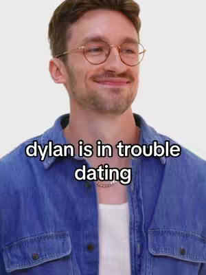 The cover image features a man with glasses and a light complexion, smiling. He is wearing a blue denim shirt over a white undershirt, and a silver chain is around his neck. In the foreground, slightly above the center, the words 'dylan is in trouble dating' are displayed in a white, sans-serif font.