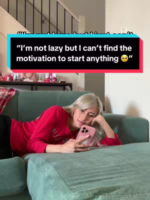 A woman lies on a green sectional sofa, resting her head on her hand while holding a pink phone. A text overlay in a black box reads, "I'm not lazy but I can't find the motivation to start anything 🥺". In the background, a stairway and decorative items are visible.