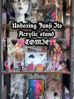 A vertical image showcases a display shelf filled with Junji Ito-themed merchandise. Bold text overlays on the image announce an unboxing of an acrylic stand. The items are arranged on wooden shelves against a backdrop which includes manga artwork. The composition has a decorative assortment of acrylic stands, artistic depictions, and thematic decorations conveying collection and presentation.