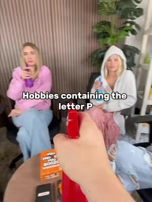 A first-person perspective image featuring three women, with elements suggesting a game or activity. The focus is on a red spray bottle held in front of the camera. Two women are visible in the background, each holding a spray bottle in a room with wood-paneled walls and a plant. The overall lighting is bright. A box of cards is on the table behind the spray bottle.