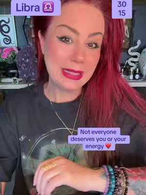 A woman with bright red hair and makeup is centered in a selfie-style shot. Behind her is a cluttered background filled with decor and a framed picture. She's speaking directly at the camera, looking animated and engaged. Below, a purple rectangle contains a white text message, with a red heart at the end. The background has a soft, diffused light, adding to the scene's intimate feel.
