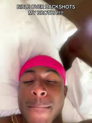The image is shot from a first-person perspective, showing a person lying in bed. The person, appearing to be a young Black man, is the central focus, with his face prominently displayed. He's wearing a pink durag and has his eyes closed. The bed appears to have a white pillow and a white sheet. The lighting seems soft, suggesting an indoor setting.