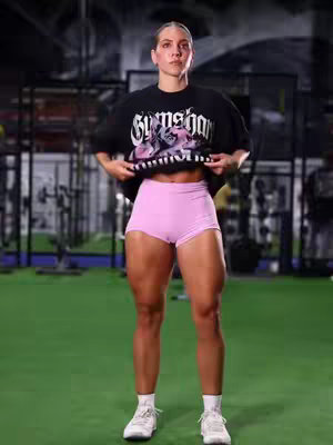 A woman stands in a gym setting, holding up her black t-shirt with the Gymshark logo. She is wearing light pink shorts and white sneakers. The background has gym equipment.