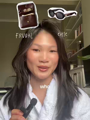 A woman with long dark hair, wearing a white robe, is centered in the frame, holding a microphone. Above her head, there are two cartoon-style images; one a brown coin purse with coins, and the other a pair of black sunglasses. Text is overlaid above each image. The background includes a blurred interior space, possibly a home.