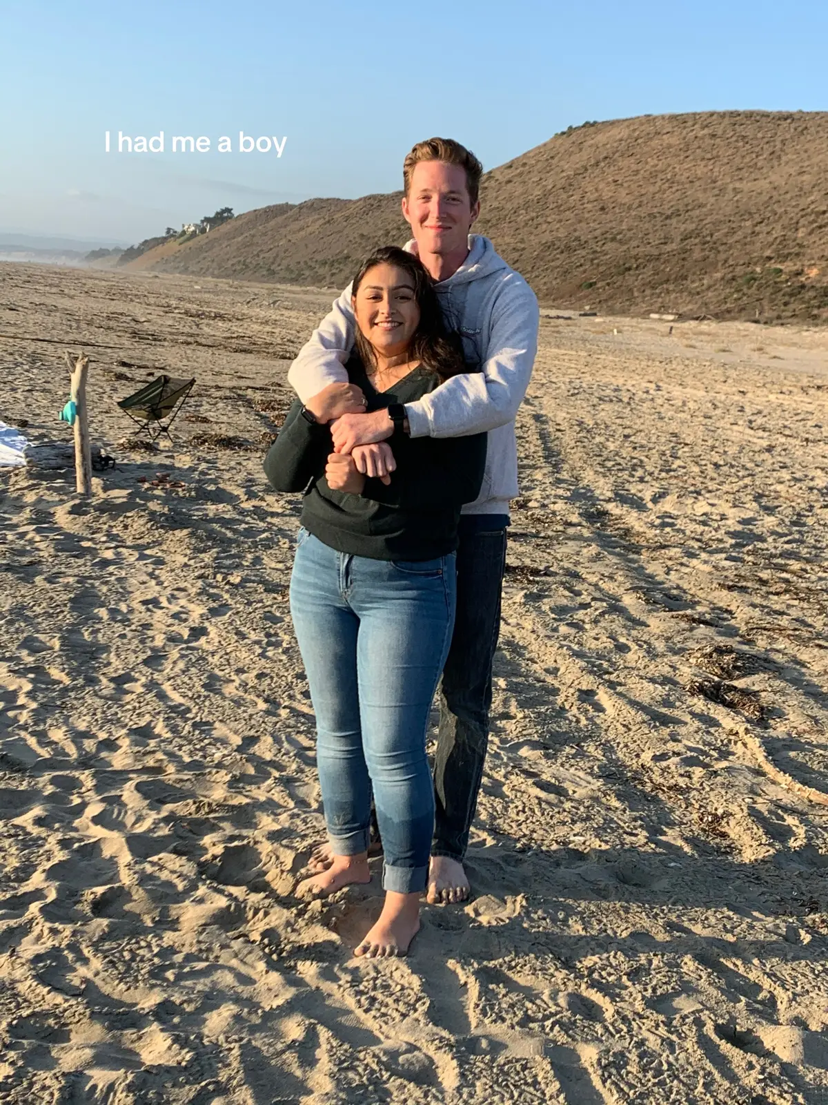 A couple embraces on a beach at sunset. The woman is in the foreground with the man behind her, hugging her. The background features a sandy beach, ocean, and hills under a clear sky.