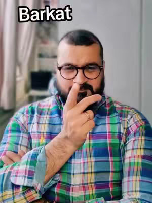 A man with a trimmed beard and glasses, wearing a colorful plaid shirt, is giving a peace sign. The image has a soft background, possibly indoors, with text overlayed at the top.