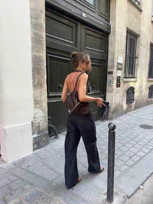 A woman in black pants and a dark top walks away from the viewer on a cobblestone street. She carries a bag and a phone, with the background of a large wooden door and a stone wall.
