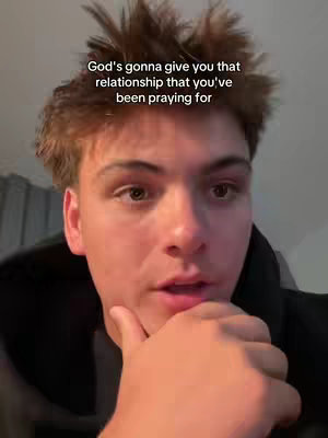A medium shot features a young man with light skin. He is looking directly at the camera with a slightly concerned expression and his right hand is resting under his chin, and the text overlay reads 'God's gonna give you that relationship that you've been praying for'. The background is blurred, with indications of an interior setting, likely a residence.