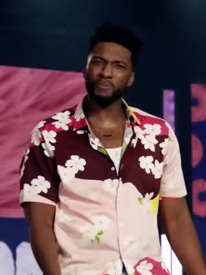 A man of color with a short, cropped haircut and a short beard is seen from the chest up. He's wearing a boldly patterned button-down shirt in light pink, burgundy, and white. The shirt has a repeating floral design. The man has a slight frown or neutral expression on his face. The background is largely out of focus, suggesting a stage or performance setting.
