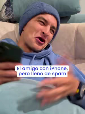 A man with blue beanie and hoodie is reacting with a face of disbelief and holding a phone while sitting on a couch. Text overlayed on the bottom: "El amigo con iPhone, pero lleno de spam".