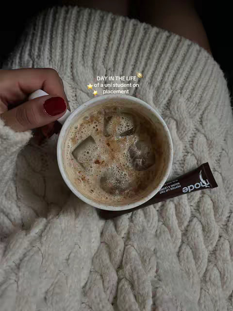 A close-up shot shows a hand holding a white mug containing a beverage with ice cubes. A dark brown lip balm is on the right. The mug and hand are on a cream-colored knit blanket. The overlay text says 'DAY IN THE LIFE of a uni student on placement'.