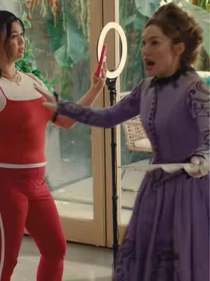 Two women in a modern setting are interacting, one dressed in contemporary athletic wear and the other in a Victorian era dress. A ring light is positioned in the center, and the women appear to be filming with a phone.