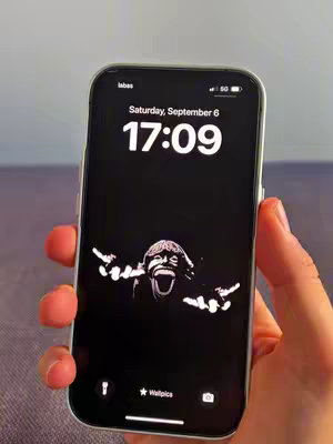 A person holding a black iPhone showing a black wallpaper featuring a white, stylized image of a face screaming with its arms raised. The image is overlaid with a digital clock that reads 17:09 and Saturday, September 6. The display is set against a background, which is potentially a fabric surface, in a slightly blurred out fashion.