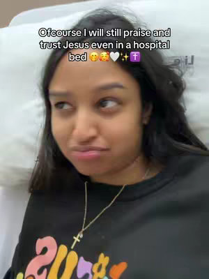 A young woman with long dark hair is lying in a hospital bed. She's wearing a black t-shirt with colorful graphic elements on it, a gold necklace, and what appears to be a cross-shaped pendant. The background is a plain white hospital setting, with a pillow supporting her head. She has a slight face expression. Text overlaid on the image reads, "Ofcourse I will still praise and trust Jesus even in a hospital bed" followed by emojis.