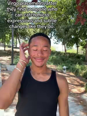 The image is a selfie-style shot of a young man outdoors. He is smiling with his eyes closed and has one hand touching his hair near his forehead. He is wearing a black tank top. The background features trees, foliage, and a glimpse of a sidewalk or path. The lighting appears to be natural sunlight.