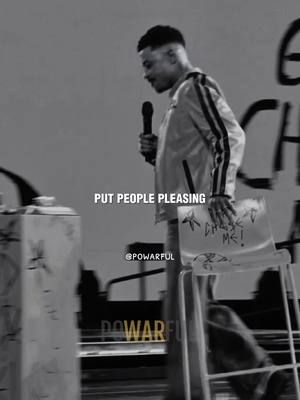 A black and white image depicts a man speaking on a stage with a microphone in his hand. Text overlayed on the image reads "PUT PEOPLE PLEASING". The man is wearing a light-colored jacket with black stripes down the sleeves. A white chair with writing on it is held under his arm. He is wearing pants and stands near a white pillar with more indistinct text on it.