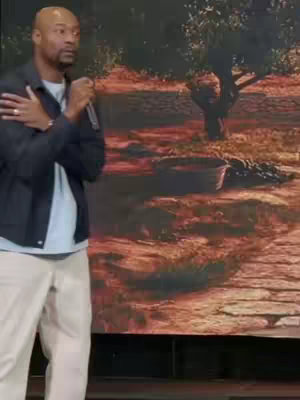 A man, wearing a black jacket over a light blue shirt and off-white pants, stands on a stage with a microphone, arms crossed. The background is a blurry landscape painting.