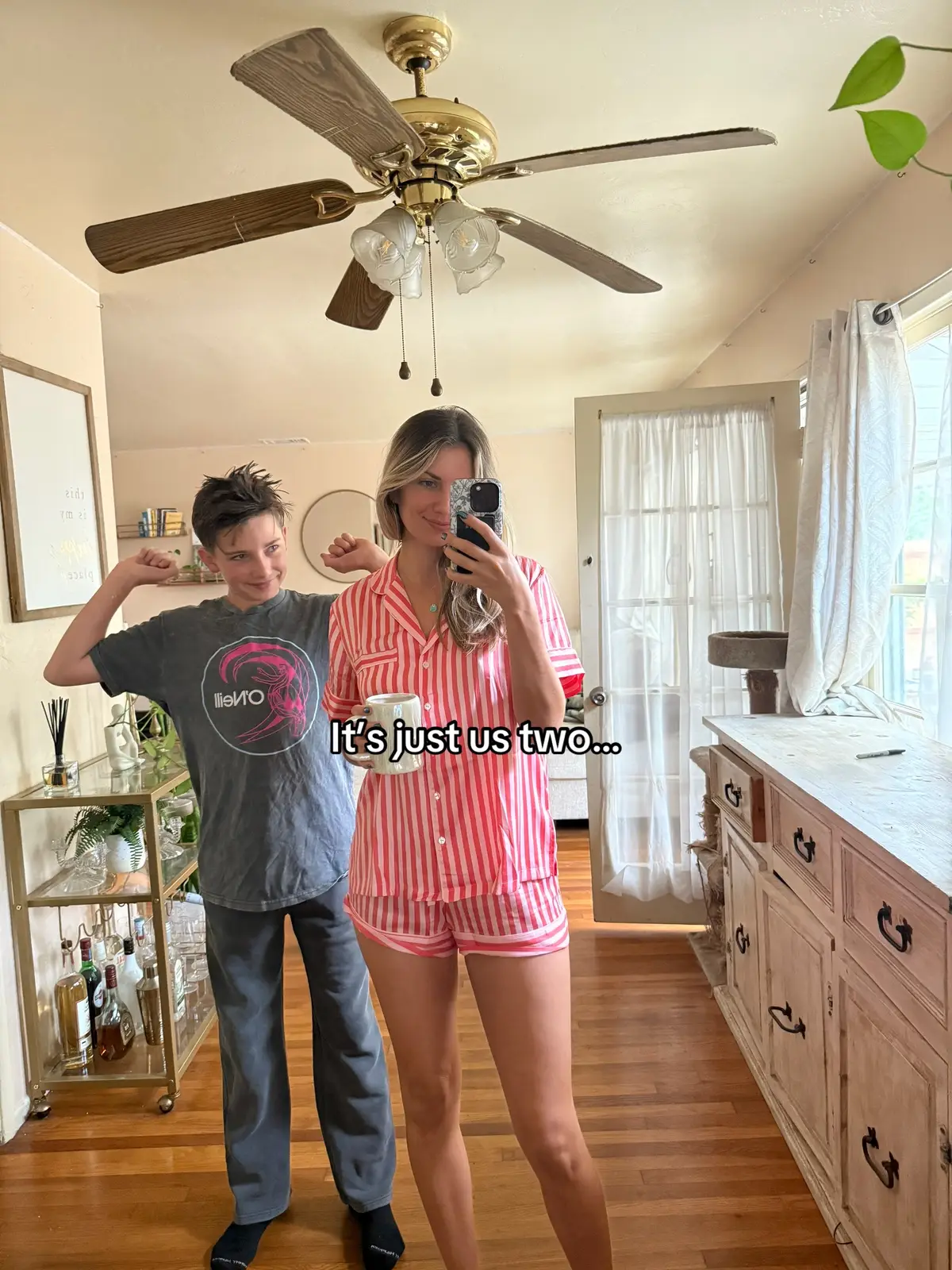 The cover image shows a woman and a young boy posing in what appears to be a home setting. The woman, wearing a red and white striped pajama set and holding a mug, is taking a selfie in a mirror. The boy, wearing a gray t-shirt and dark pants, is flexing his arms. The interior has a warm color palette, with a wooden floor, a large ceiling fan, and a decorative vanity.