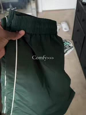 A person's hand holding a green track pant with a white stripe and the word Comfy>>> overlaid.