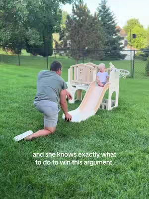 A man in a green shirt kneels on the grass, appearing to interact with a small slide set up on a lawn. A baby is seated at the top of the slide. The setting is a well-kept yard with a fence in the background.