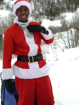 The cover image features a male, identified as NBA Youngboy, dressed in a Santa Claus costume. He is standing in what appears to be a snowy environment. He's wearing a red Santa suit with white trim, a Santa hat, and black gloves. He has a gold chain. The background is out of focus and mountainous.