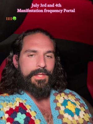 A man with long, dark hair and a full beard is centered in the frame, wearing a colorful blue, yellow, red, and white patterned sweater or cardigan. The background is split with a darker upper section and the red of a car interior toward the bottom. Text overlays show the date, a spiritual concept, and a shamrock with the number 1111.