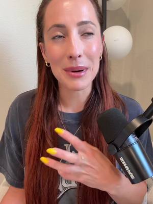 A medium shot featuring a woman with long auburn hair and a dark grey t-shirt, is positioned slightly left of center, speaking into a microphone with a serious expression. Her left hand gestures toward the right with yellow-painted fingernails. A black microphone occupies the lower right corner. The background is a neutral off-white.