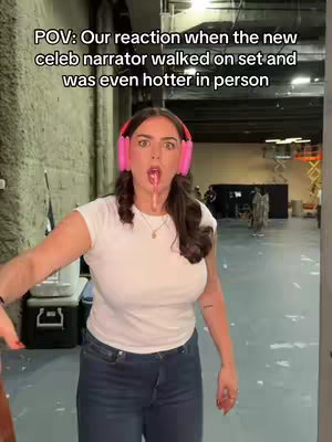 A woman stands in a doorway inside a large studio or warehouse-style space, mid-reaction. She's wearing a white t-shirt, jeans, and large pink headphones. Her mouth is open, and a clear, thin, waxy substance appears to be dripping from it. The background shows industrial details such as lighting rigs, equipment, and some people standing in the distance, suggesting a film or video set. The lighting is bright. The woman’s expression is one of exaggerated surprise or shock.