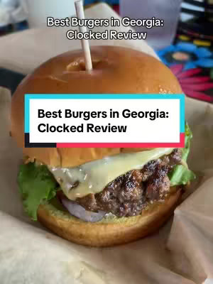 A close-up shot of a burger with cheese, lettuce, a visible onion ring. The burger is on a cardboard wrapper with text overlays. The background shows more blurred food items, and is set mostly in brown hues.