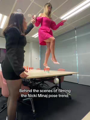 Two women are in an office setting. One is balancing on a table while the other appears to be assisting, possibly capturing a trending pose. Cups are stacked on the table. The background is an office environment with windows, a beige table, and minimal decorative items. The woman on the table wears a pink dress, while the other wears a black outfit.