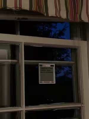 The image shows a window with a sign posted on it. The window is divided into panes and has a patterned valance at the top. The sign is in a frame and appears to be handwritten.