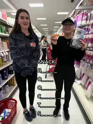 The cover image shows two young women in a brightly lit store, standing in a shopping aisle. The woman on the left is wearing casual outerwear and black leggings. The other woman, positioned to the right, is dressed in all black and holds a bag of Chain Link snacks. Text overlay creates a fill-in-the-blank game with "SNACK" above, which is partly filled out, suggesting audience interaction.