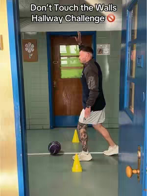 A man prepares to throw a ball in a hallway. The image is framed through a doorway. The man is wearing athletic clothing, there are cones and a ball set up as though he is about to play a game or do an exercise. Text at the top says, “Don’t Touch the Walls. Hallway Challenge”.
