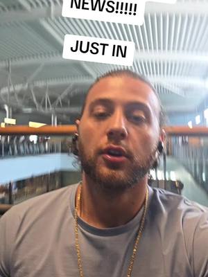 A man with a short beard and light-colored hair looks toward the camera. He is wearing a blue shirt and gold chain. Two white text boxes with black text overlay the image, reading "NEWS!!!!!" and "JUST IN". The background is blurry and shows an indoor setting with metal bars and structural supports.