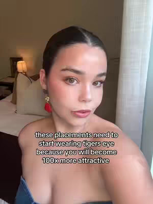 A woman with fair skin and dark hair looks directly at the camera. She is wearing minimal makeup, with blushed cheeks and a subtle lip color. Red earrings are visible. The background is softly blurred, with a bed and window curtains. Text overlay indicates a video about wearing tiger's eye.