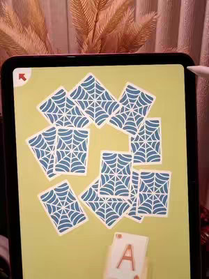 The cover image shows a tablet displaying a game of spider solitaire. The game board is a yellow background with a pile of playing cards, each featuring a blue spiderweb design. The playing cards are arranged in a loose, scattered fashion. In the foreground, a single Ace card is visibly displayed. The overall composition is simple, with the focus on the gameplay. The background is a blurred view of pink and brown.