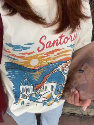 The image shows a person wearing a white t-shirt with a printed graphic of a seaside scene with the word 'Santorini' on it. The person is holding a bag, and the background suggests an urban or college setting. The lighting seems natural, possibly daylight.