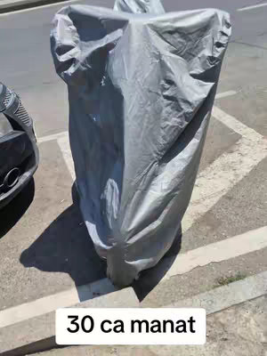 An outdoor shot of a covered motorcycle. The motorcycle is covered with a gray, reflective tarp. The tarp's shape suggests the form of a motorcycle. The background is blurred, showing a sidewalk and part of a vehicle. A text overlay at the bottom of the frame reads "30 ca manat" in a white rectangular box.
